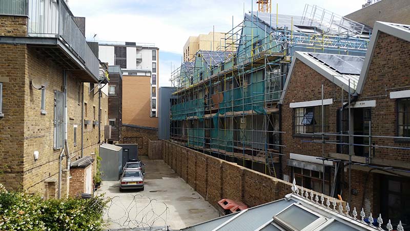 A narrow alleyway between brick buildings, with parked cars on the left and a construction site with scaffolding on the right, showcases an Essex architect’s work beside a historic party wall under a partly cloudy sky.