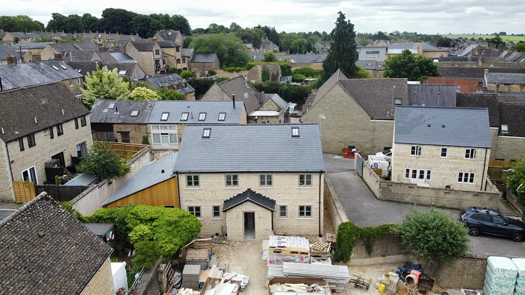 Aerial view of a residential neighborhood with stone houses; a newly constructed house, possibly designed by an Essex architect, features building materials in the front and is central in the image.
