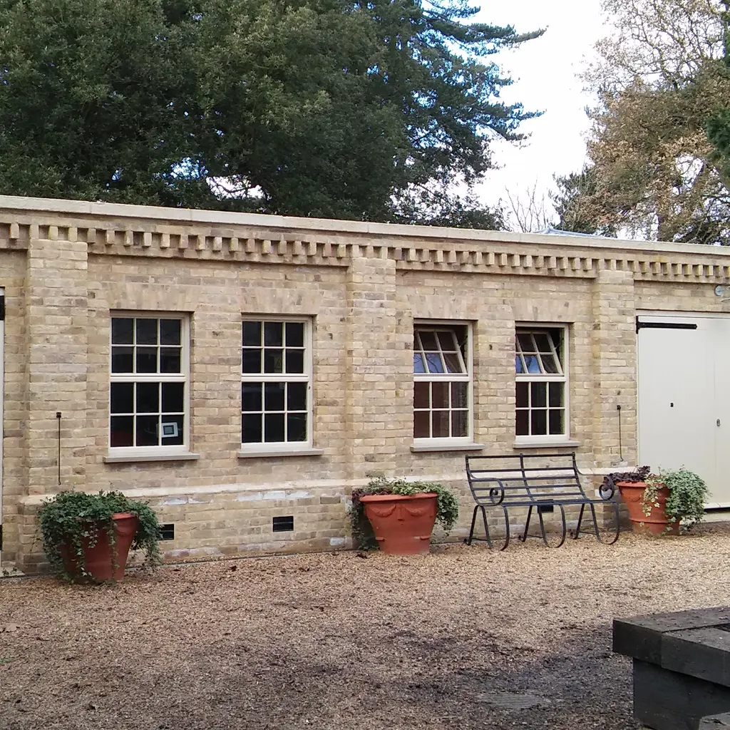 Single-story brick building in Essex with three windows, large white double door, gravel ground, potted plants, and a black metal bench in front. Designed by an architect, the property features trees in the background.