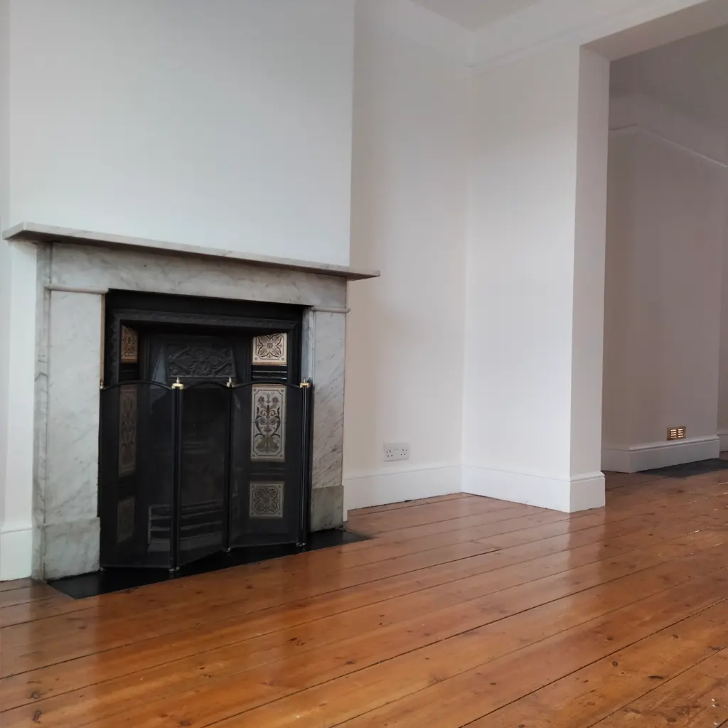 A fireplace with a decorative metal screen and marble surround, designed by an Essex architect, is set against a white wall in a room with polished wooden floors.