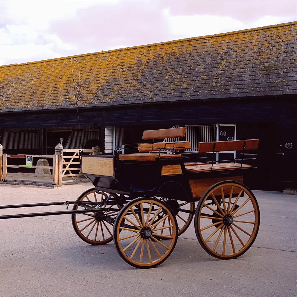 A vintage wooden horse-drawn carriage with large spoked wheels stands on a paved area in front of a rustic barn, reminiscent of classic Essex countryside scenes admired by any architect.