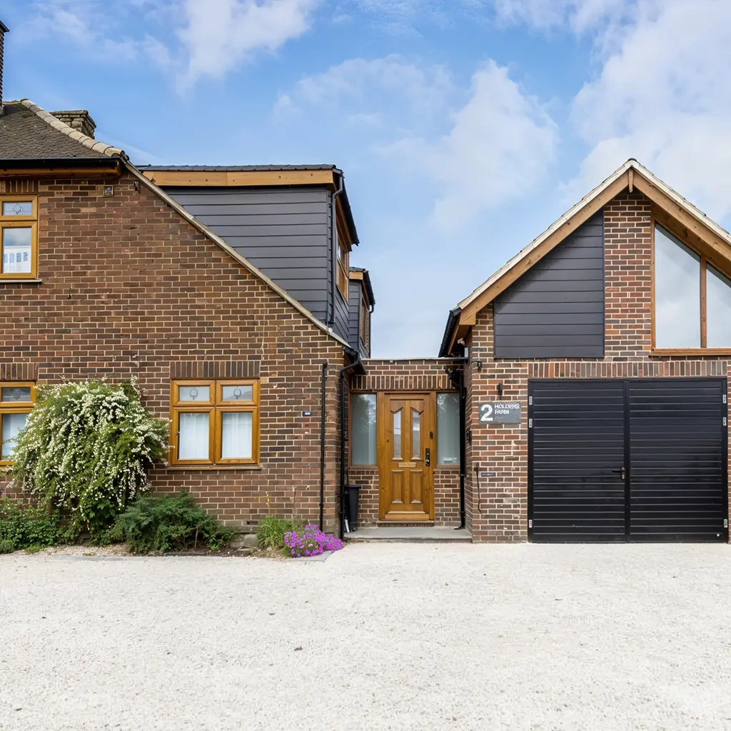 A modern brick house in Essex with dark siding, a brown front door, large windows, a black garage door, and a gravel driveway under a partly cloudy sky—expertly designed by an architect with attention to party wall details.