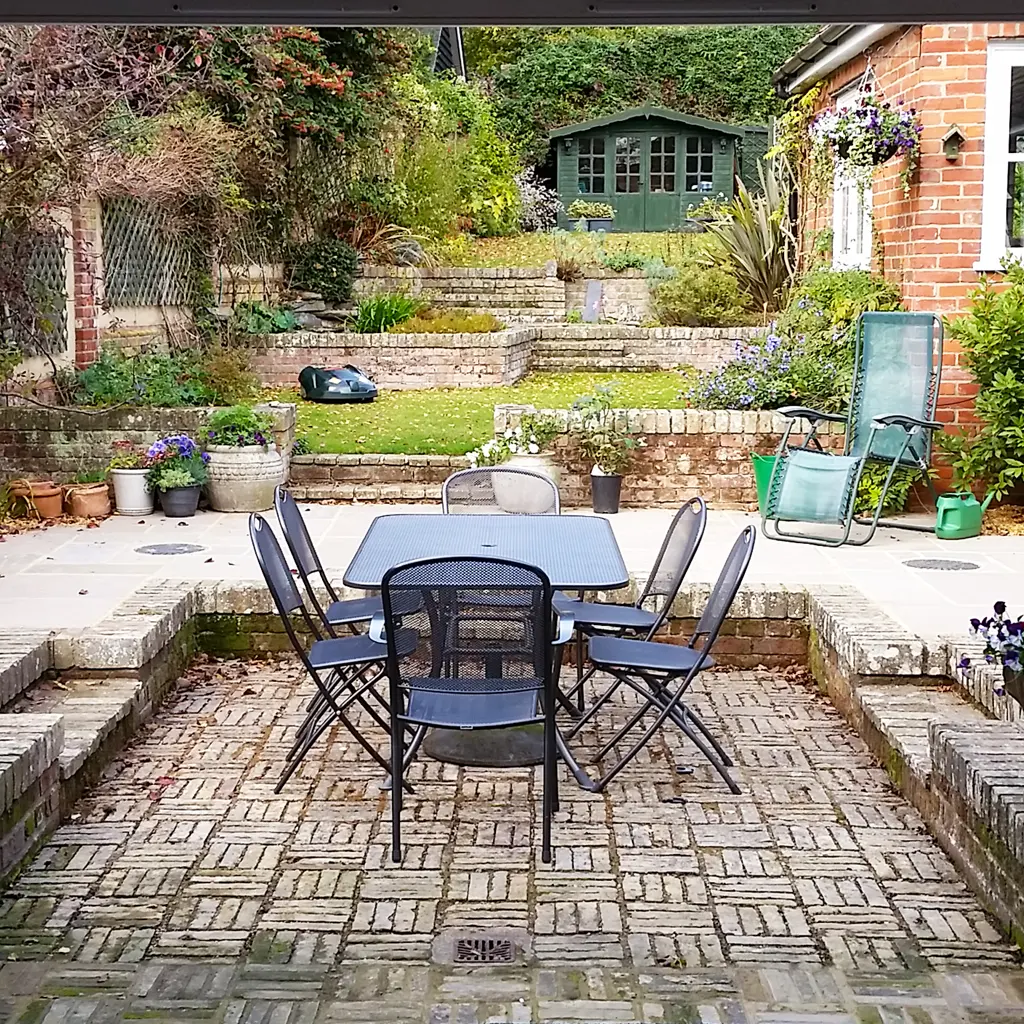 A patio with a square table and six chairs is set in a brick courtyard, beside a classic Essex party wall, surrounded by lush garden plants, steps, and a green shed in the background.