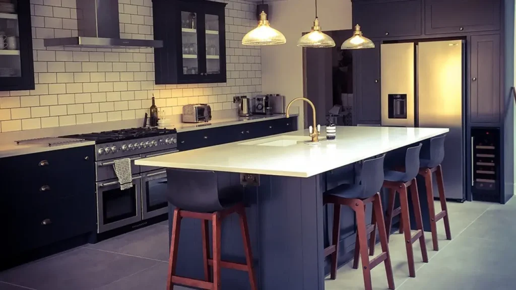 Modern kitchen with dark cabinets, white subway tile backsplash, stainless steel appliances, an island with a white countertop designed by an Essex architect, four bar stools, and pendant lights overhead.