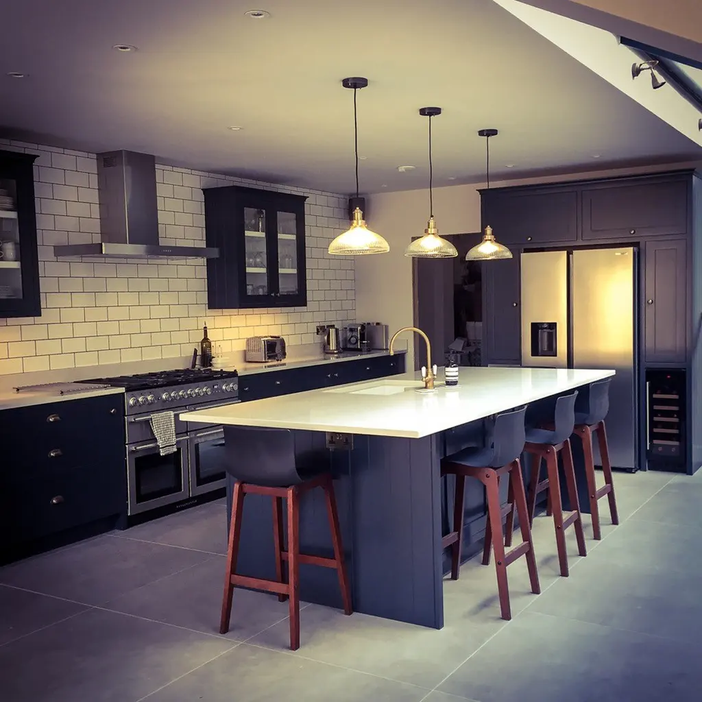 Modern kitchen designed by an Essex architect, featuring dark cabinets, white subway tile backsplash, large island with white countertop, four bar stools, pendant lights, and sleek stainless steel appliances.