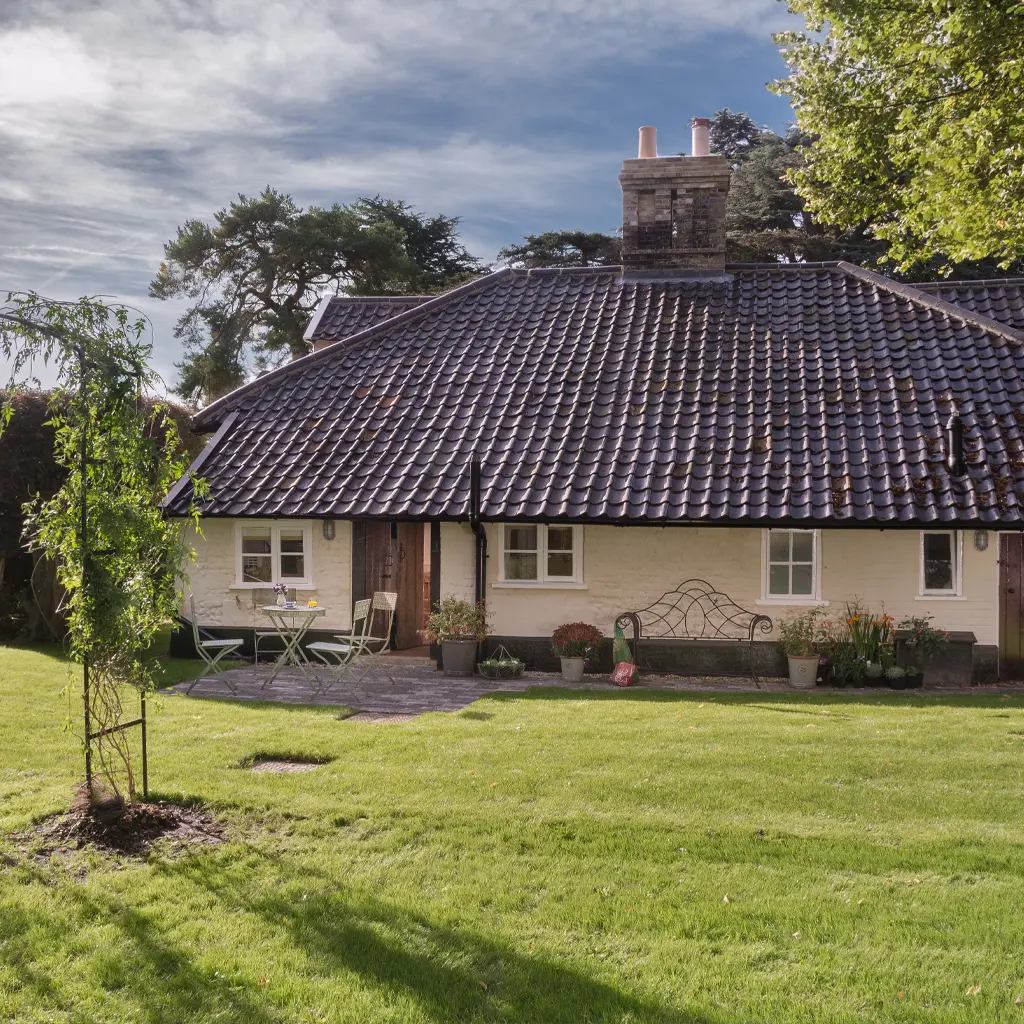 A small cottage with a dark tiled roof and cream-colored walls sits on a well-kept lawn in Essex, featuring outdoor seating, potted plants, and trees in the background.