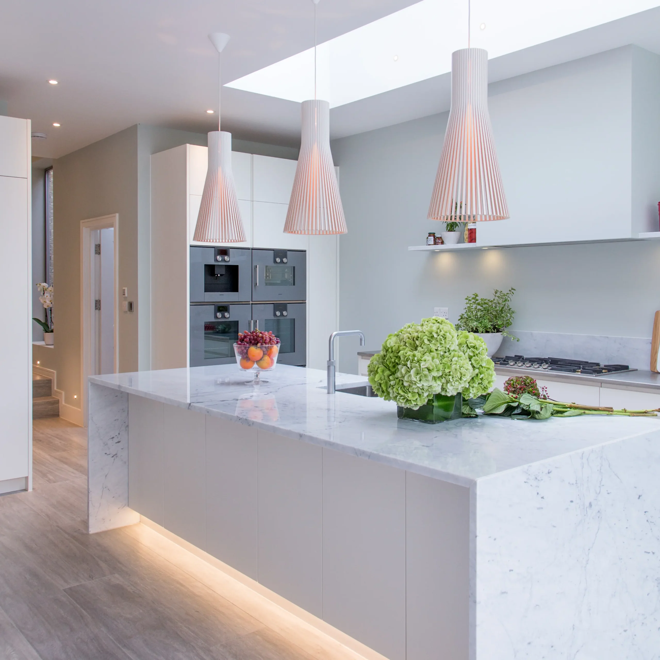 Modern Essex kitchen with a marble island, green hydrangeas, pendant lights, built-in ovens, and light wood flooring. Designed by an architect, it features fruit bowls and kitchen essentials on the counters.