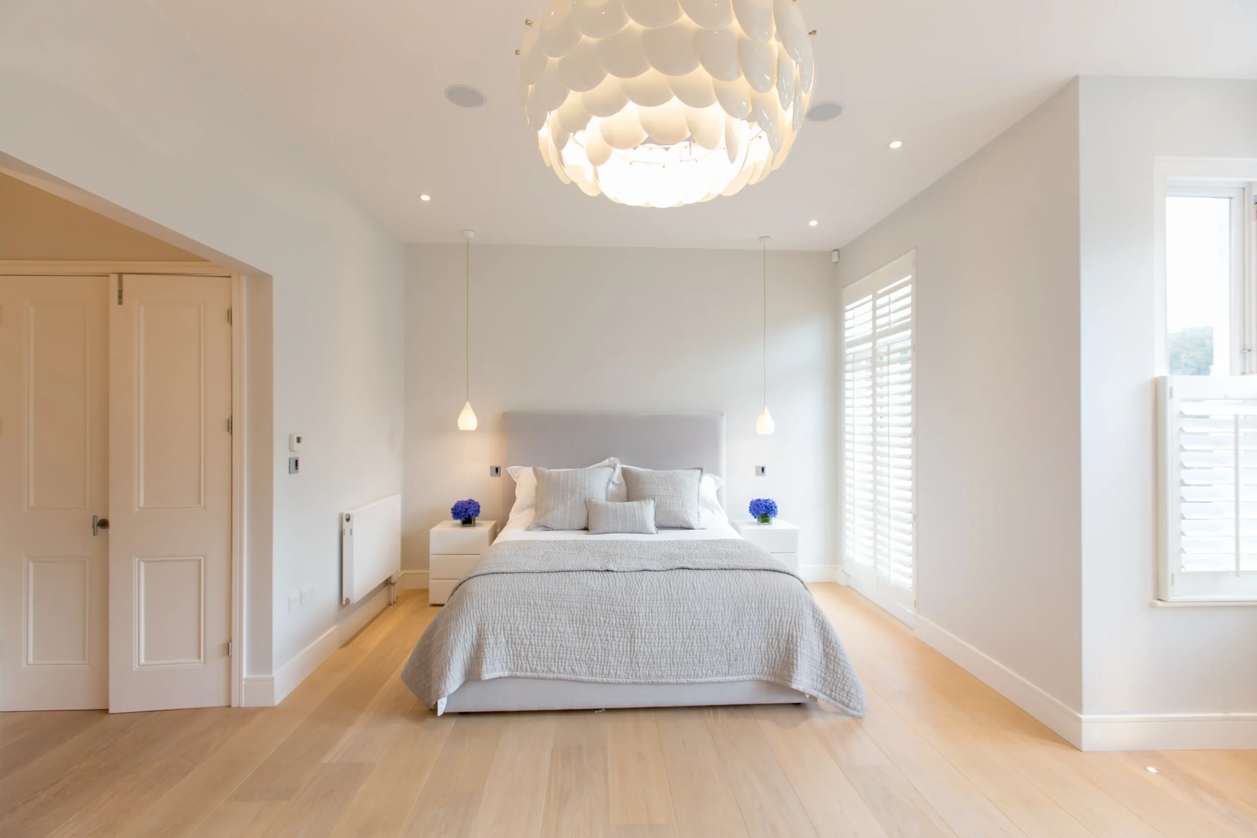 A modern, bright bedroom designed by an Essex architect, featuring a large bed with white bedding, two pendant lights, nightstands with blue flowers, and a striking decorative ceiling light.