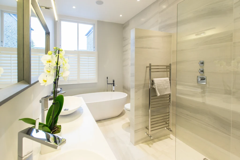 Modern bathroom with a freestanding bathtub, walk-in shower, double sink, heated towel rail, and an orchid plant on the counter—expertly designed by an Essex architect, all in a bright, neutral color scheme.