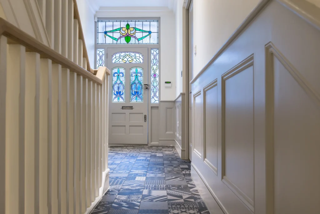A hallway in Essex with patterned tile flooring, white paneled walls, a wooden staircase, and a white front door featuring colorful stained glass panels reflects an architect’s eye for detail.