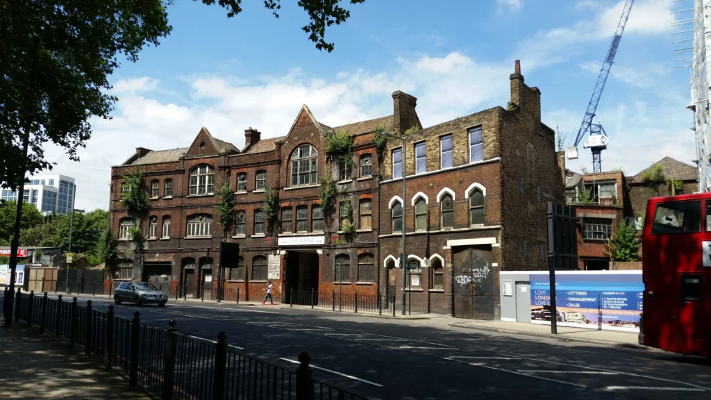 A large, old brick building with boarded windows and visible decay—perhaps once designed by a famed architect—sits empty beside a street in Essex, where a passing car trails behind a red double-decker bus.