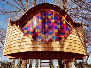 Wooden treehouse with a curved exterior and a large, colorful stained glass window in the center, designed by an Essex architect, supported by tree trunks, with leafless trees in the background.