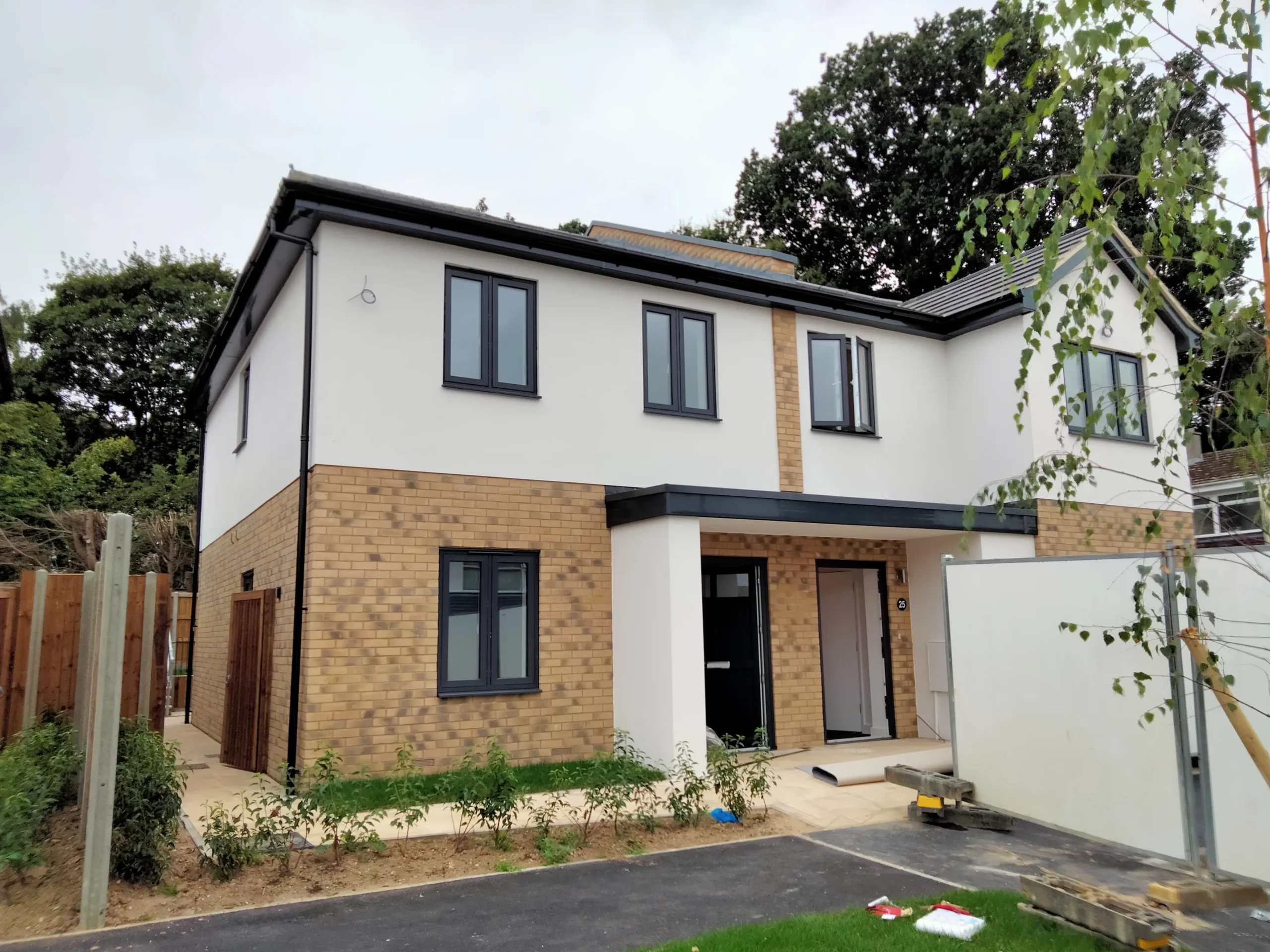 Two-story modern semi-detached house in Essex with a brick and white exterior, black-framed windows, a small front garden, and construction materials nearby—designed by a skilled architect with attention to the party wall features.