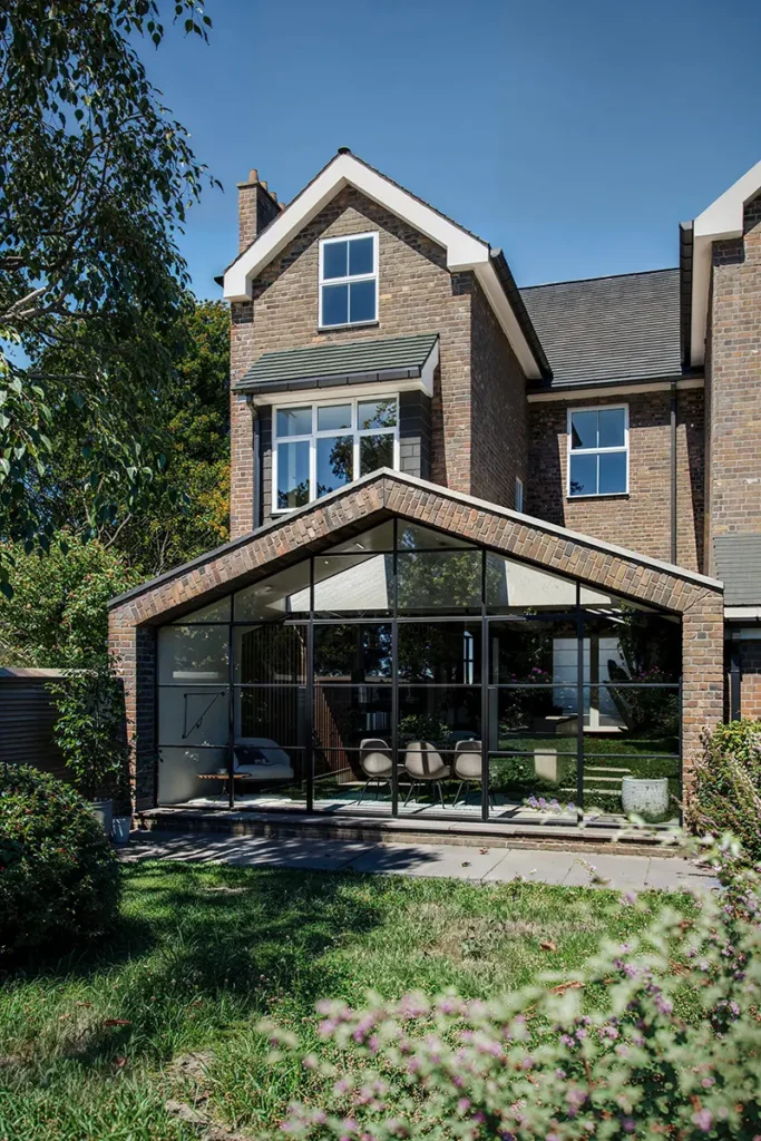 A modern brick house in Essex with large glass windows and a glass-enclosed extension, designed by a skilled architect, surrounded by greenery and a grassy yard.