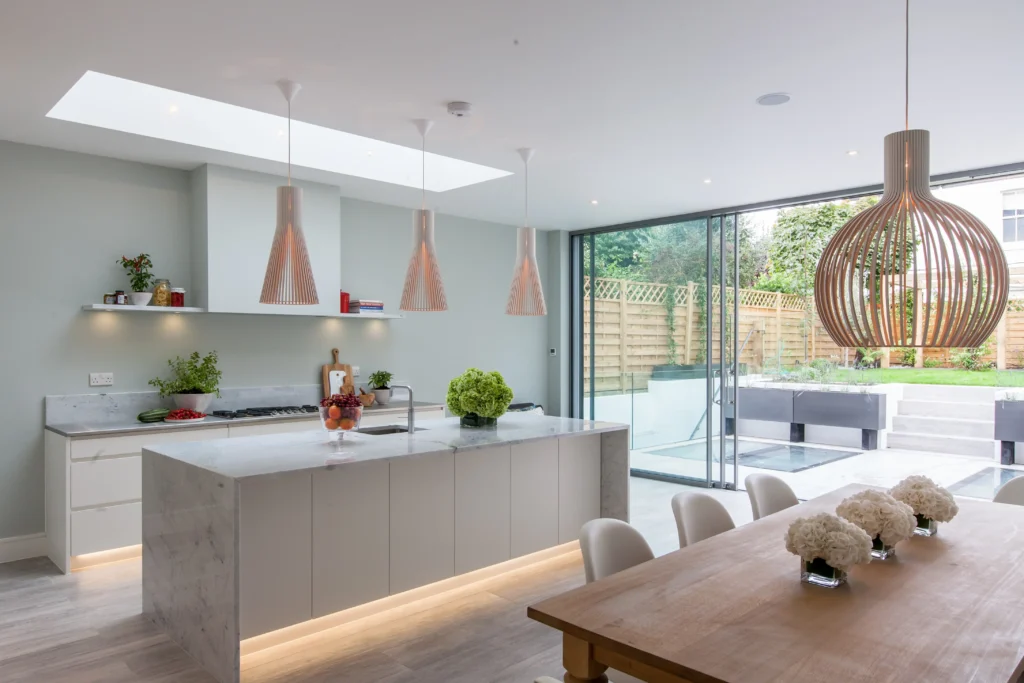 Modern kitchen and dining area in Essex, designed by an architect, featuring a marble island, pendant lights, large glass doors to a patio, and light wood furnishings.