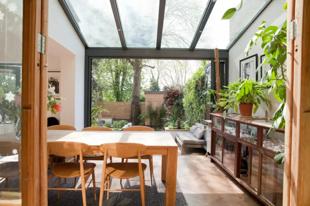 A bright dining area with a wooden table and chairs, designed by a skilled Essex architect, is surrounded by large glass windows and doors that overlook a green garden filled with various plants and trees.