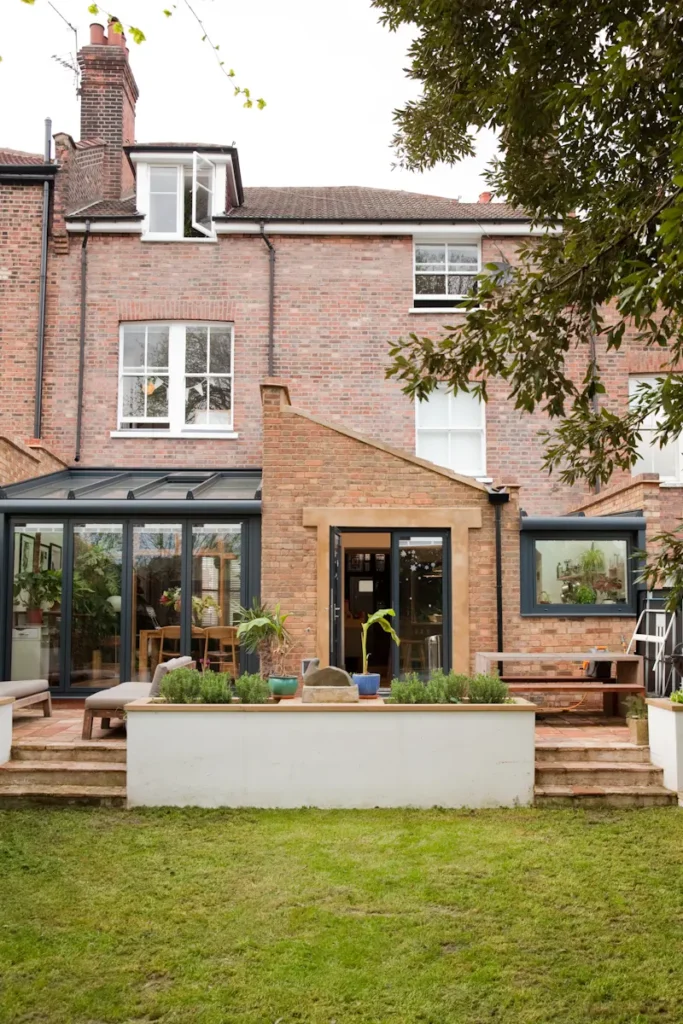 Rear view of a brick townhouse with large windows, a glass extension designed by an Essex architect, patio area, potted plants, and a grassy lawn.