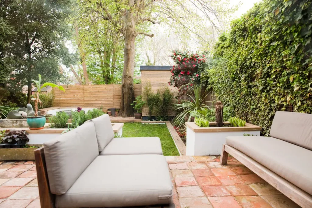 Modern backyard garden in Essex designed by an architect, featuring two cushioned outdoor sofas, tiled patio, manicured lawn, potted plants, and raised beds surrounded by trees and greenery near a classic party wall.
