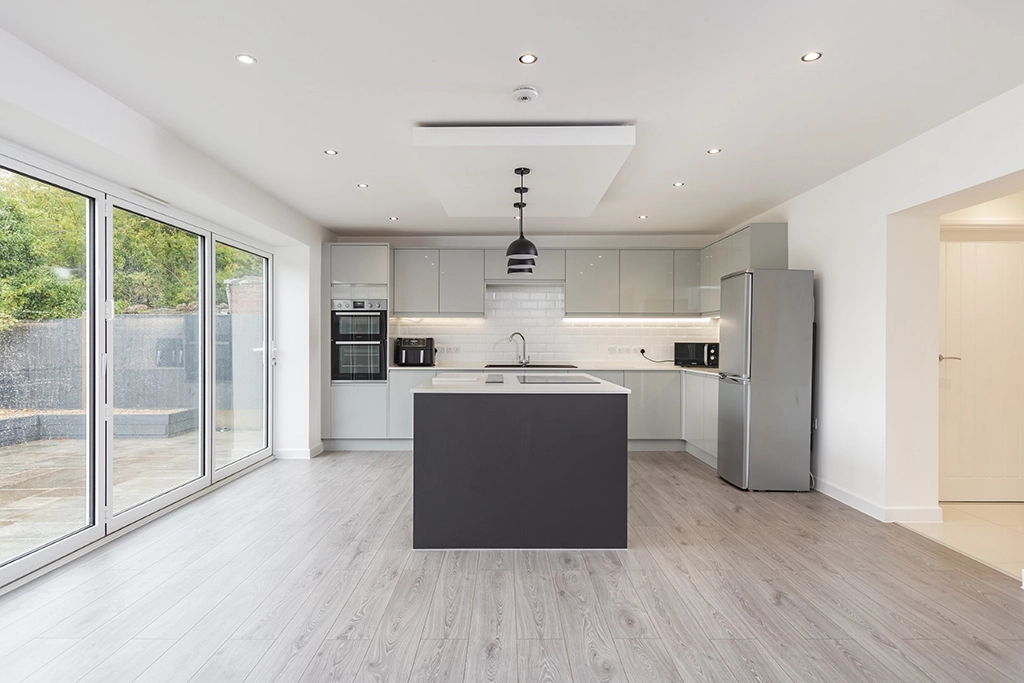 Modern kitchen with grey cabinets, a central island, built-in appliances, a stainless steel fridge, and large sliding glass doors designed by an Essex architect—perfect for entertaining or relaxing by the patio.