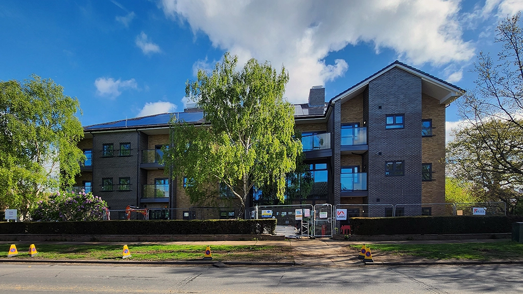 A modern three-story apartment building in Essex with large windows and balconies, partially obscured by trees under a blue sky with scattered clouds. Construction barriers highlight ongoing work on the party wall in front.