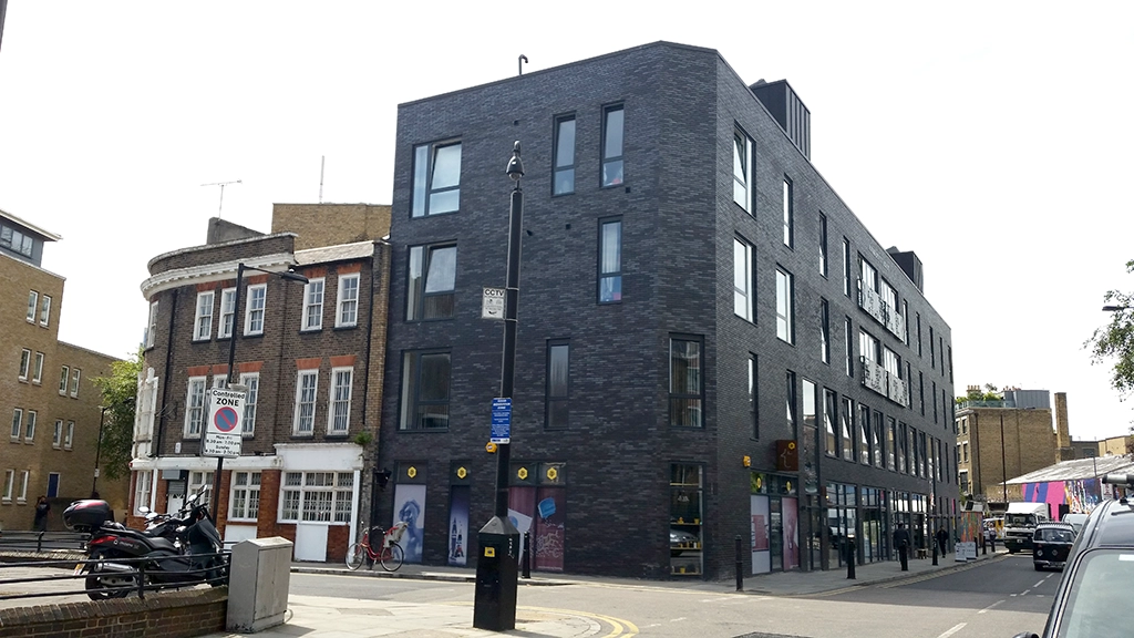 A modern black brick building designed by an Essex architect stands at a street corner next to an older red brick building with white trim; parked bikes and motorcycles line the sidewalk along their party wall.