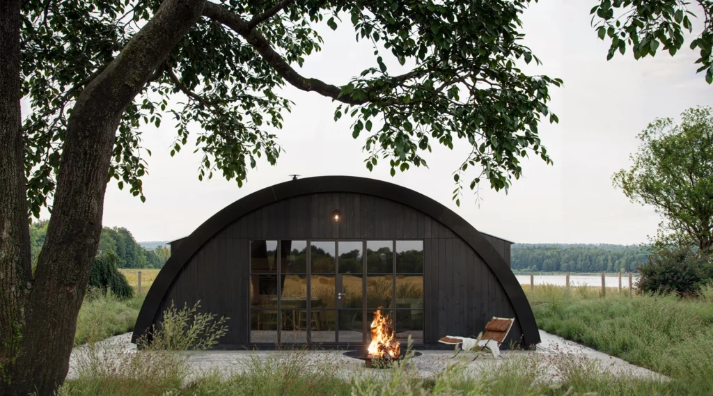 A modern black arched cabin, designed by an Essex architect, features glass doors and stands by a fire pit amid tall grass, trees, and a scenic lake in the background.