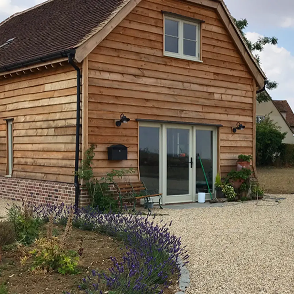 A wooden house with a brick foundation and gravel driveway, designed by an Essex architect, featuring a glass sliding door, outdoor bench, and potted plants by the entrance.