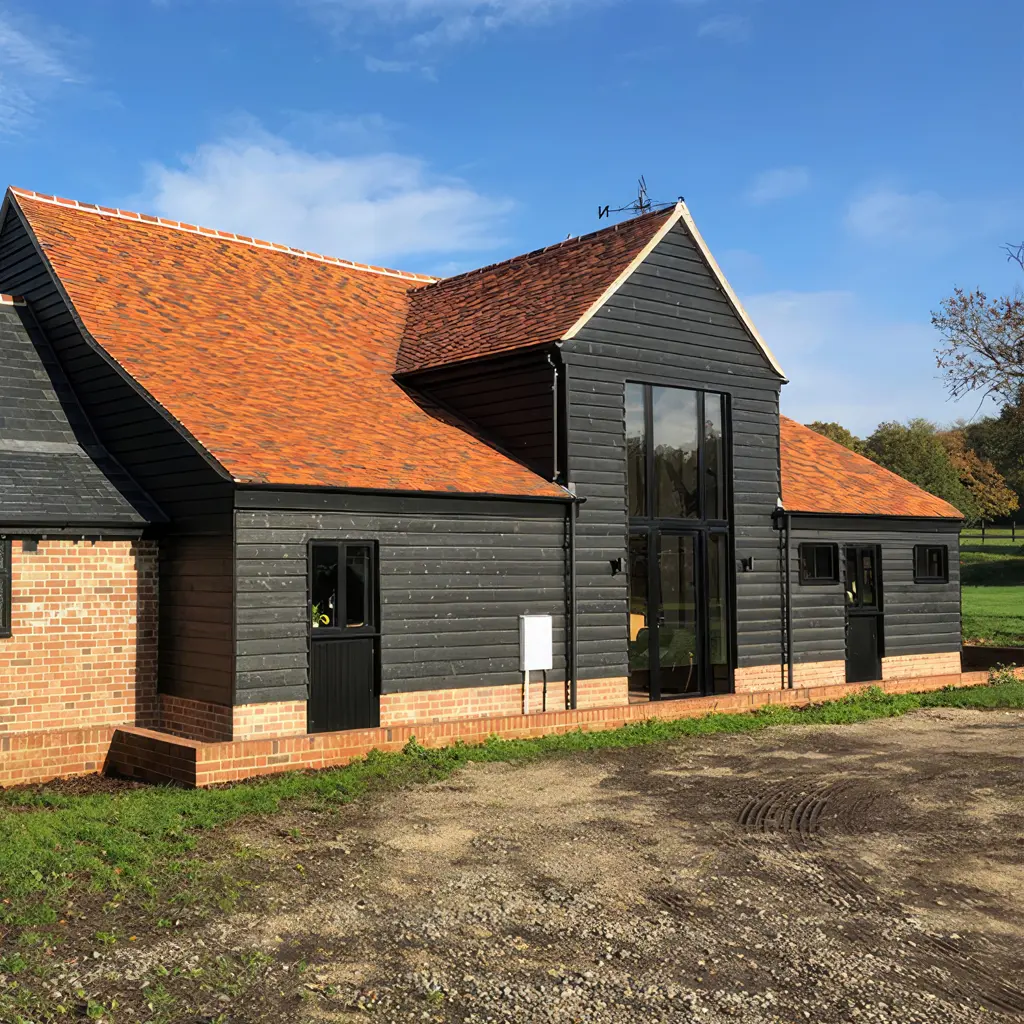 Modern barn-style building with black wooden siding, large glass entrance, orange tiled roof, and brick foundation, designed by an Essex architect, set in a rural landscape under a blue sky.