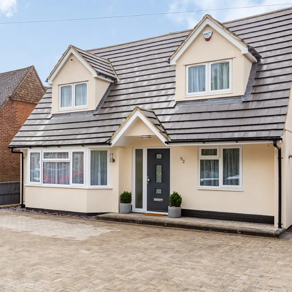 A modern detached house in Essex with cream-colored walls, gray roof tiles, dormer windows, and a dark front door designed by an architect, featuring two potted plants by the entrance.