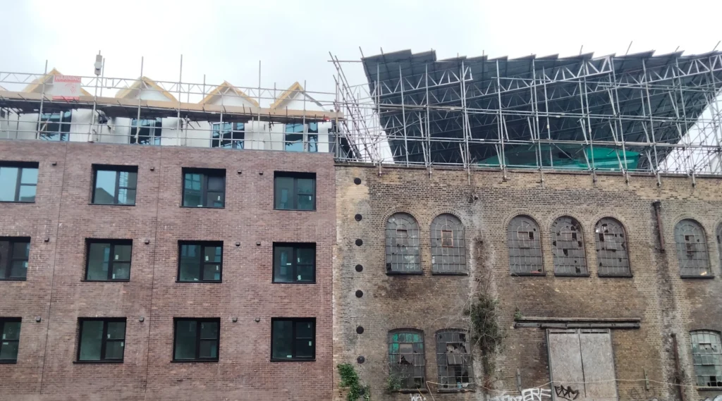 A modern brick building stands next to an older, weathered Essex structure with arched windows and scaffolding on the roof, showcasing careful architect planning along the shared party wall.