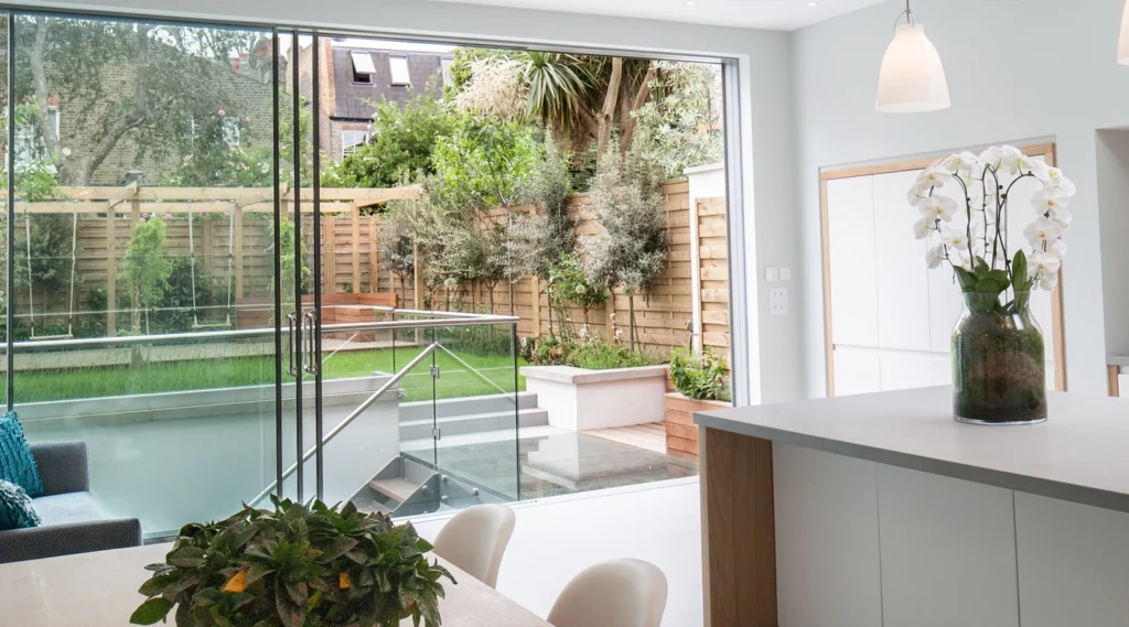 Modern kitchen designed by an Essex architect features large glass doors opening to a landscaped backyard with lawn, plants, and stairs. A white orchid in a vase graces the kitchen island.