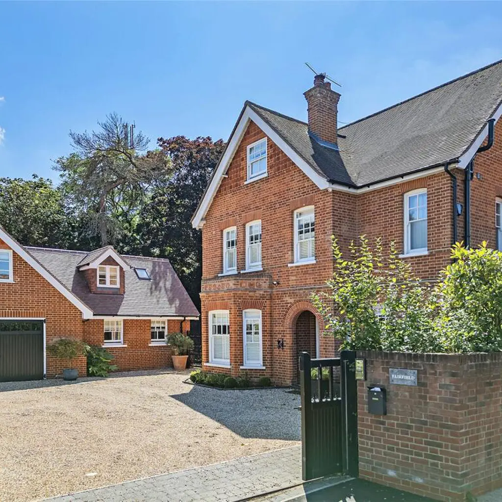 A two-story red brick house with white trim, an arched doorway, and dormer windows—designed by an Essex architect—stands next to a detached garage, behind a black gate on a sunny day.