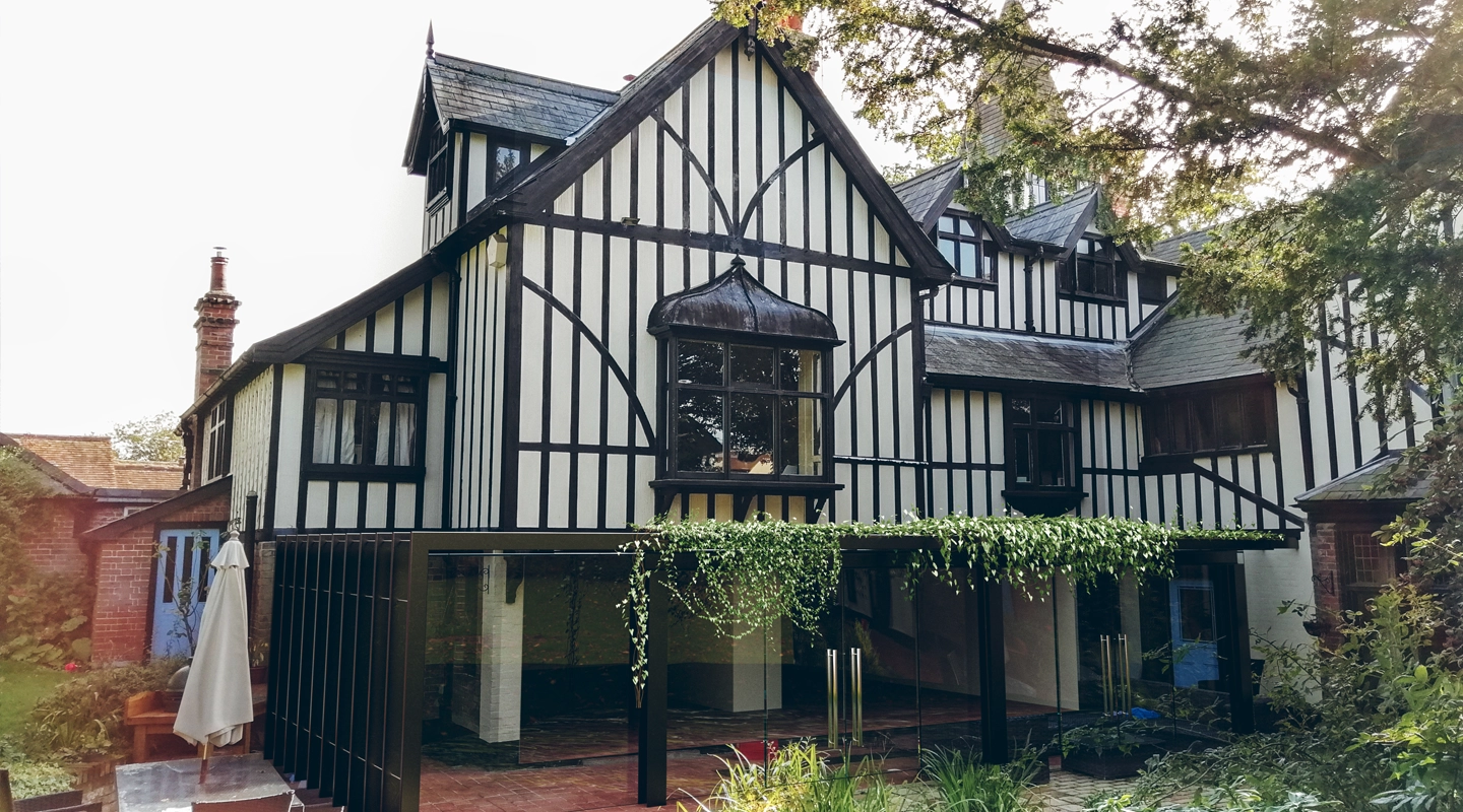 Large Tudor-style house with black and white timber framing, gabled roofs, multiple windows, and a covered patio area with hanging greenery—designed by an Essex architect, perfect for homes with a shared party wall.