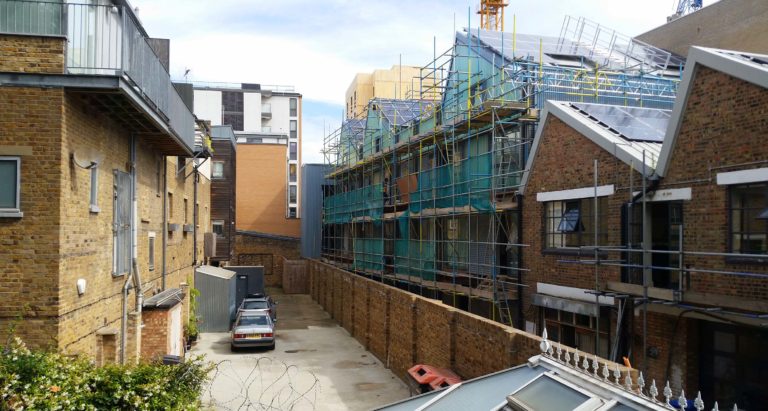 A narrow alleyway bordered by brick buildings in Essex, with scaffolding and construction work—perhaps involving a party wall—visible on the right; a parked car sits at the end of the alley.