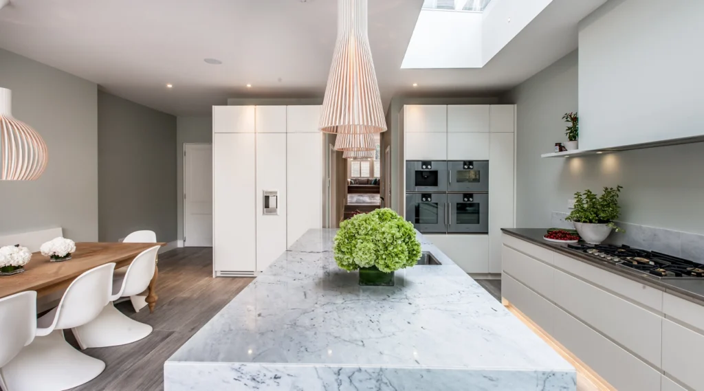 Modern kitchen with marble island, integrated appliances, white cabinets, dining table with white chairs, and pendant lighting. Designed by an Essex architect, green plants and flowers add decorative touches.