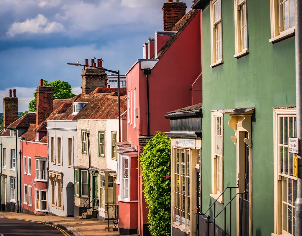 A row of colorful, old-fashioned terraced houses lines a quiet, slightly curved street under a partly cloudy Essex sky, each home thoughtfully designed by an architect mindful of every party wall.