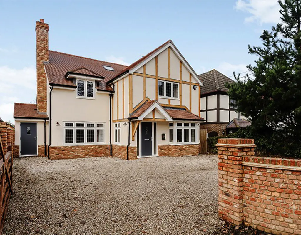 Two-story house with brick and beige exterior, designed by an Essex architect, featuring multiple windows, a pitched roof, gravel driveway, and party wall, all surrounded by a low brick wall and trees.