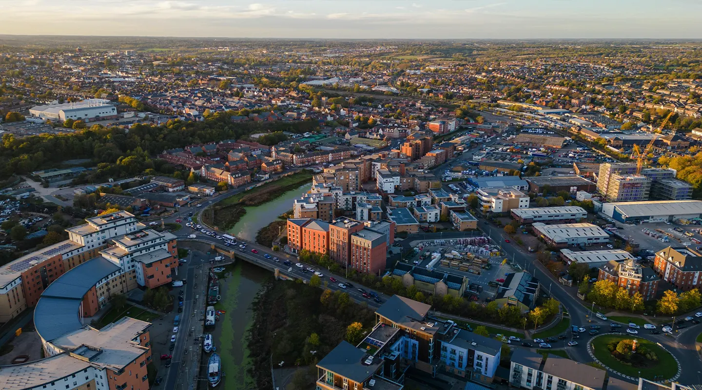 Aerial view of an Essex town with a river running through the center, surrounded by residential and commercial buildings designed by local architects, roads, and green areas under a clear sky.