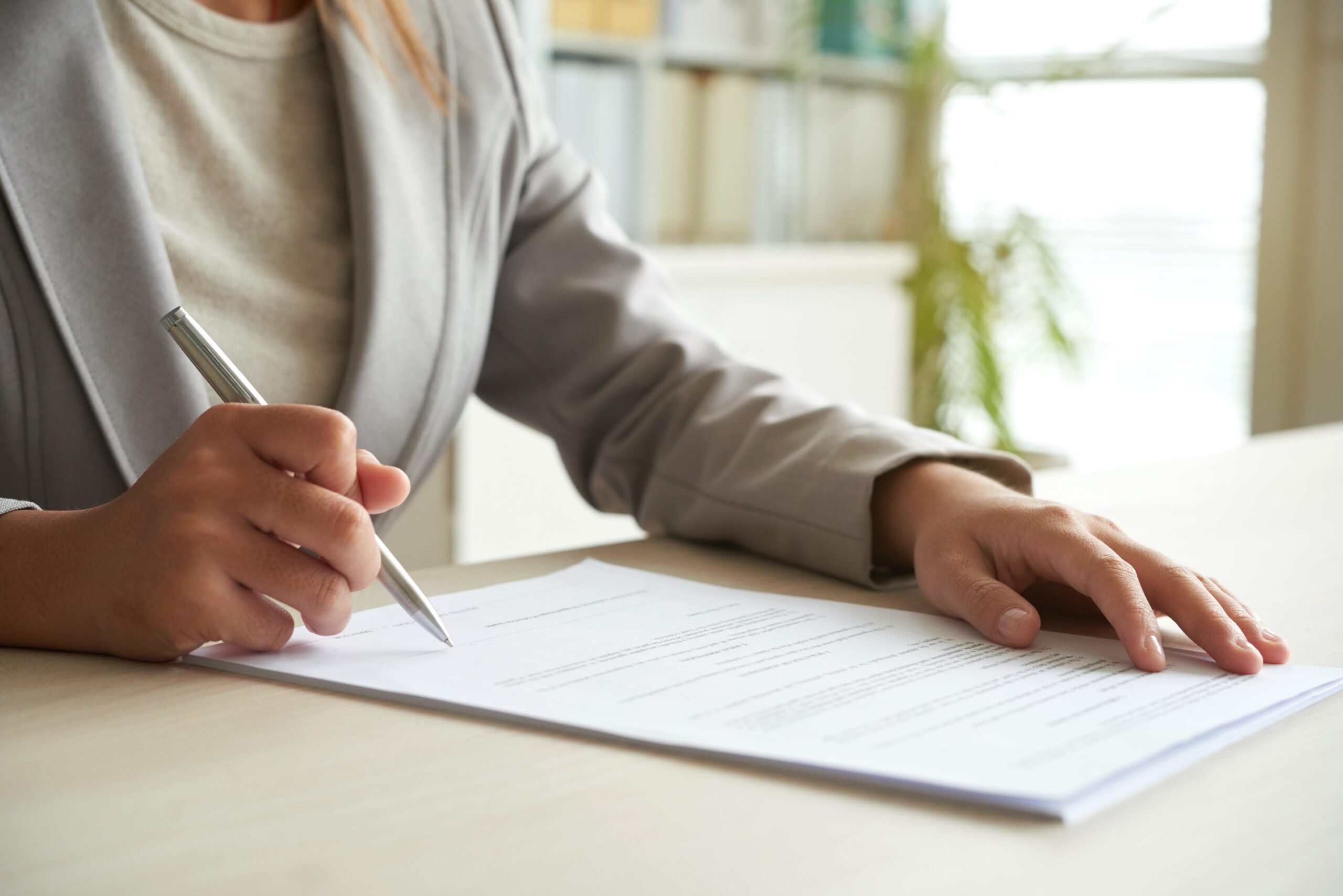 A person in a gray suit is holding a pen and reviewing or signing a document on a desk.