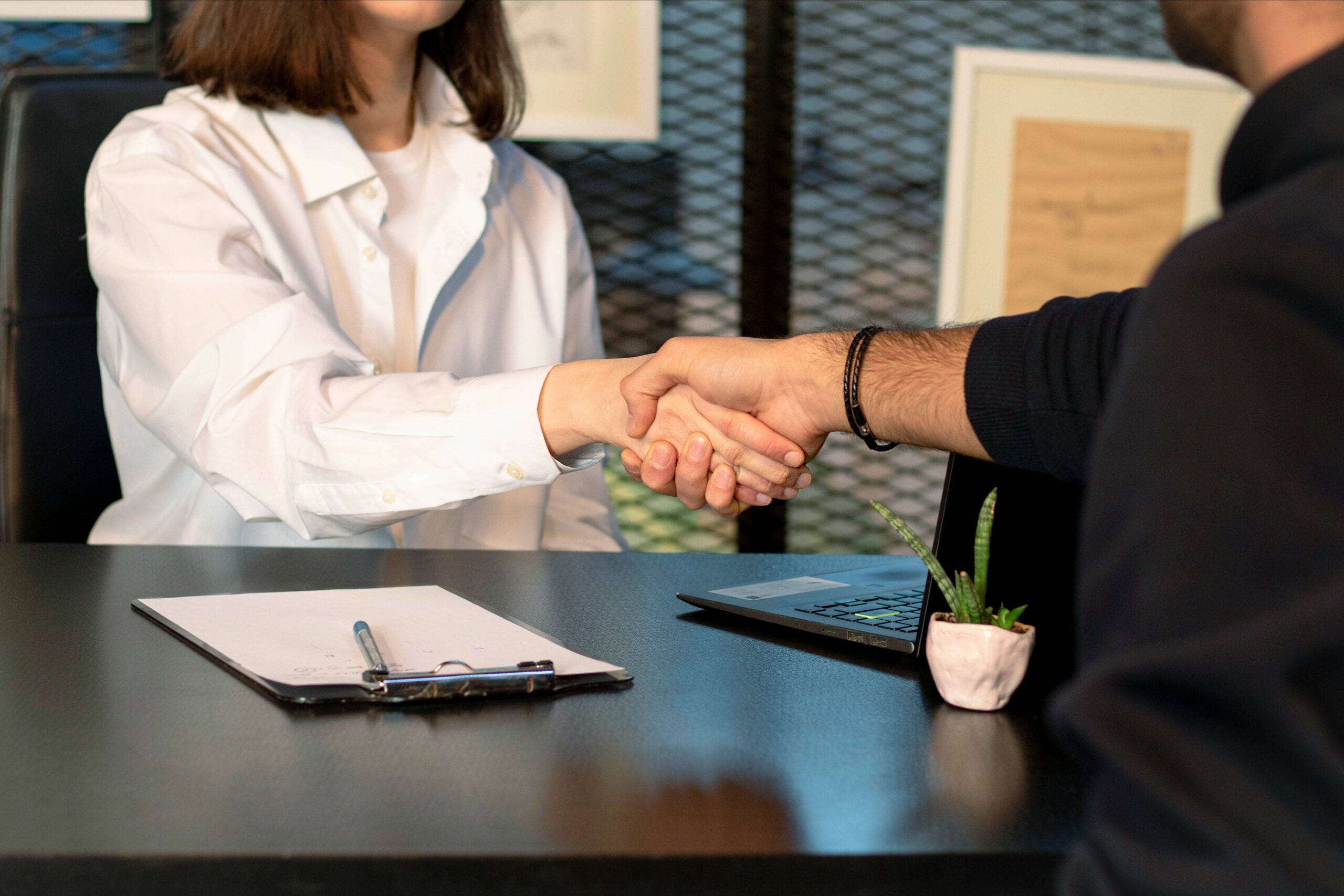 Two people sit across a desk shaking hands. A clipboard, pen, and small plant are on the desk. Only their torsos and arms are visible.