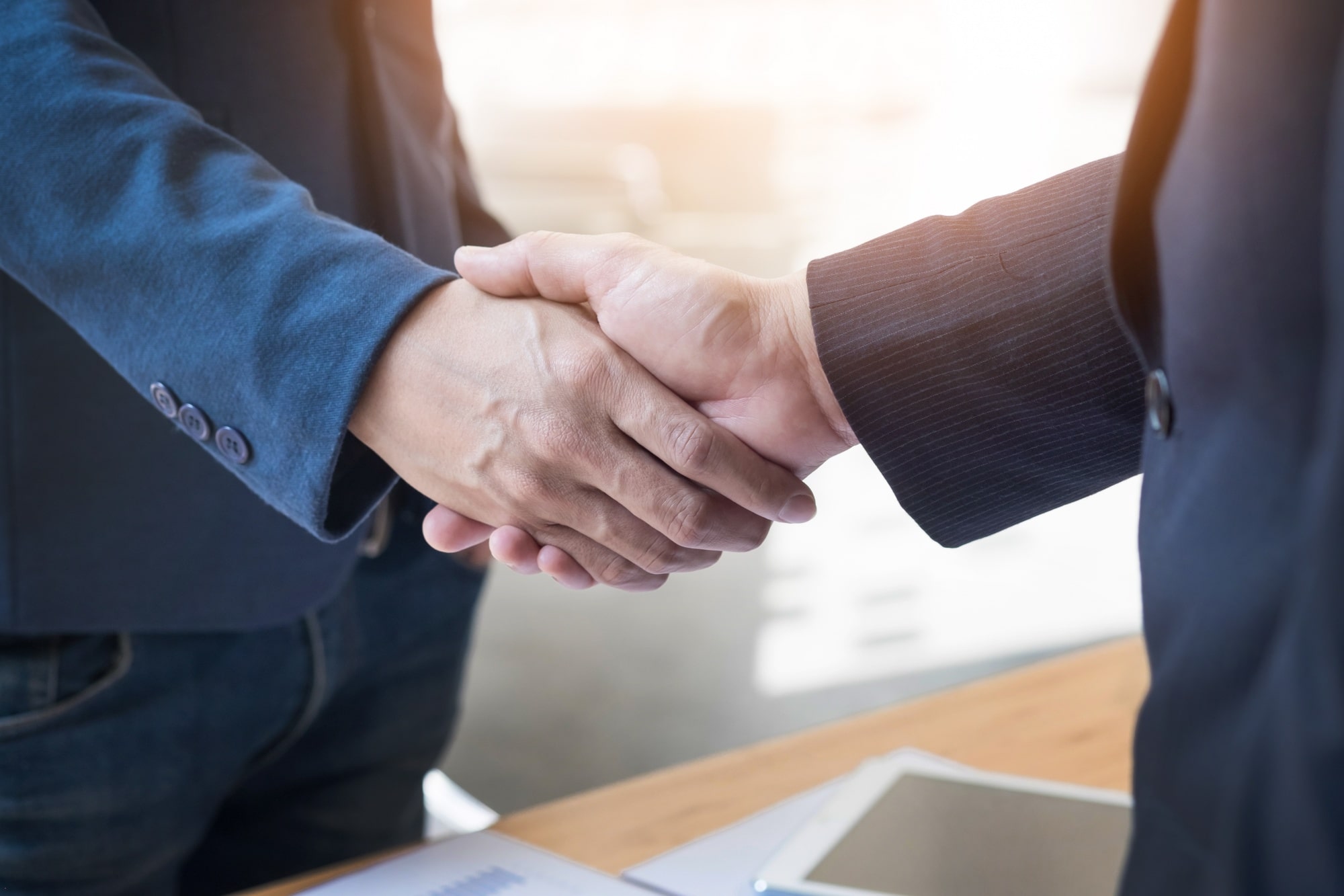 Two people in business attire shake hands over a desk with documents and a tablet, suggesting a professional agreement or partnership, possibly involving a party wall surveyor.