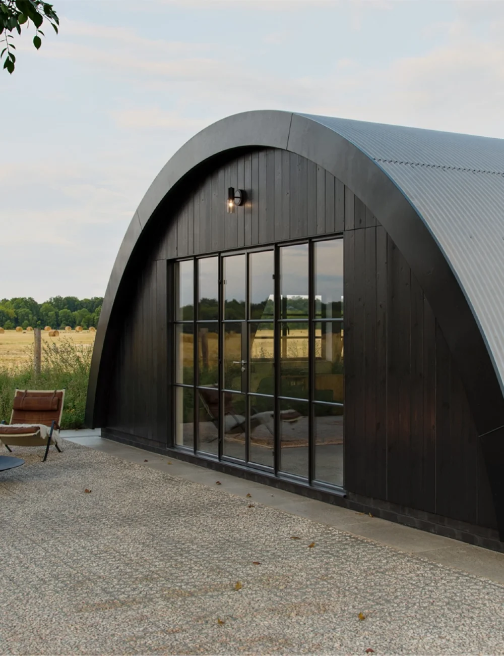 CLIENTS A modern arched cabin designed by an Essex architect features large windows and sits on a gravel patio next to a fire pit and chair, overlooking a field and trees.