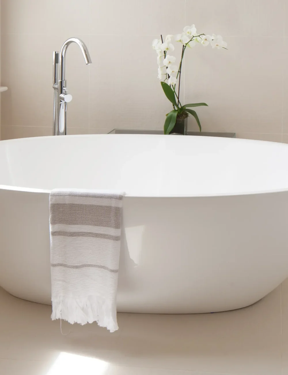IMG_7720 A modern white freestanding bathtub with a towel draped over the side, designed by an Essex architect, features a chrome faucet and a potted white orchid on a small table in the background.