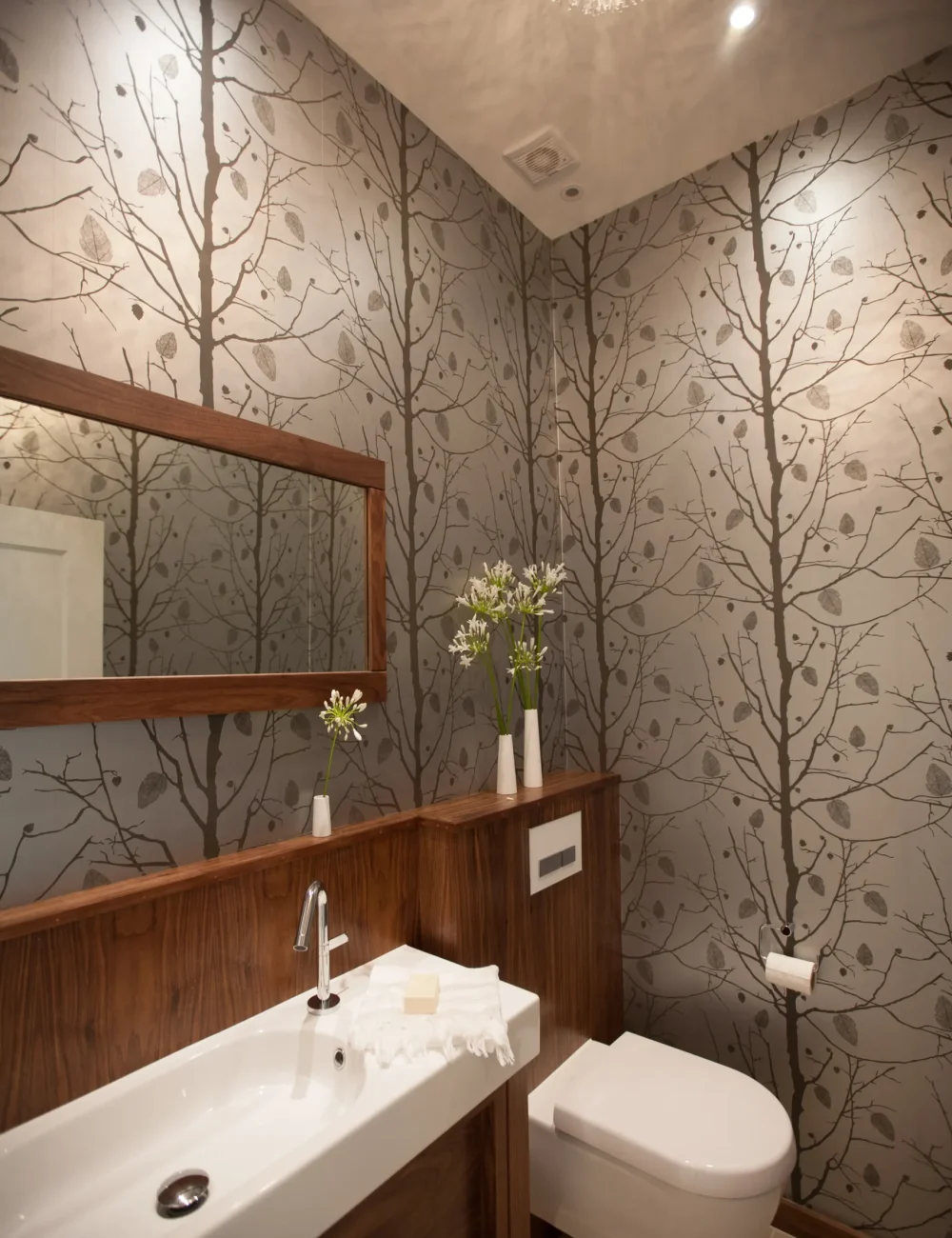 IMG_7754 Modern bathroom with wood accents, a wall-mounted sink, a large mirror, floral vases, a toilet, and tree-patterned wallpaper designed by an Essex architect on a party wall.
